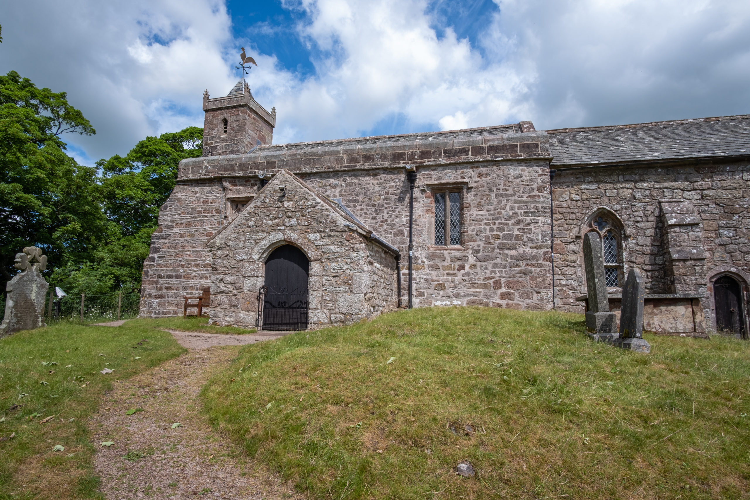 Ancient stone parish church with square tower and rooster weather vane, built on a grassy hill above the village. Old gravestones stand in the foreground beside a dirt path leading to the dark arched door. Blue sky with white clouds and green trees frame the scene.